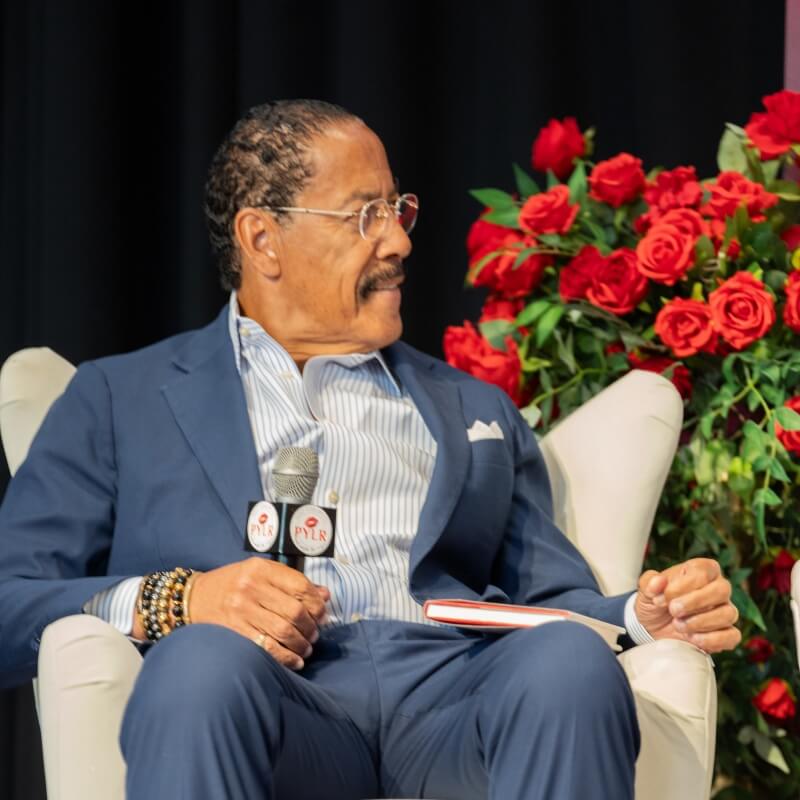 Dr. Dennis Kimbro Sitting in chair on stage at event with microphone